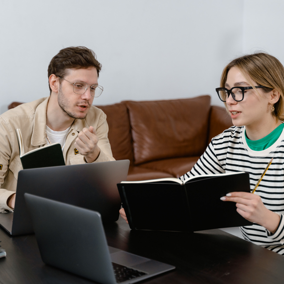 Man in beige jacket sits next to woman in black and white striped to, both in front of laptops on a black table