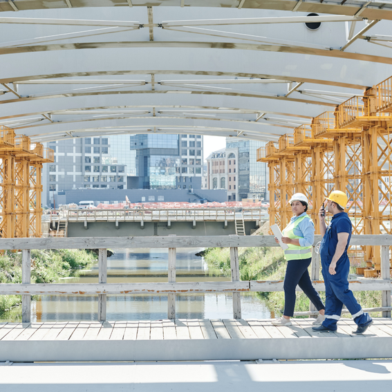 A man and a woman in hard hats walking across a walkway in a construction site