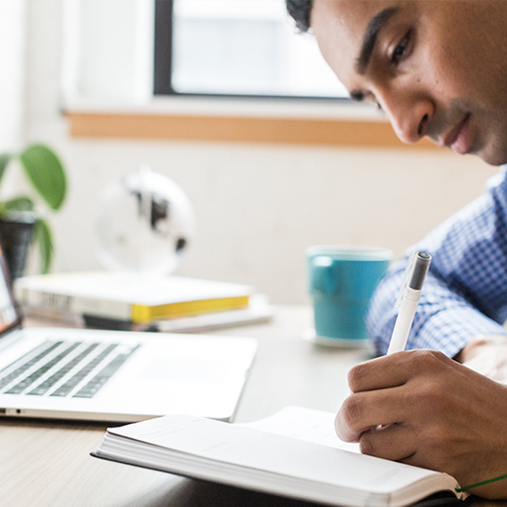 Man in shirt writing on paperwork with laptop in the background