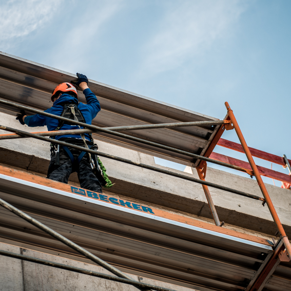 Man in blue hooded jacket and a orange hard hat stands on scaffolding