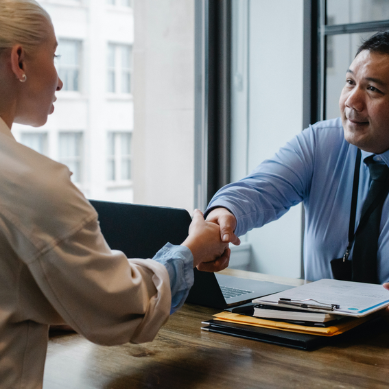 Woman in white top shakes man in shirt and tie's hand over a wooden desk